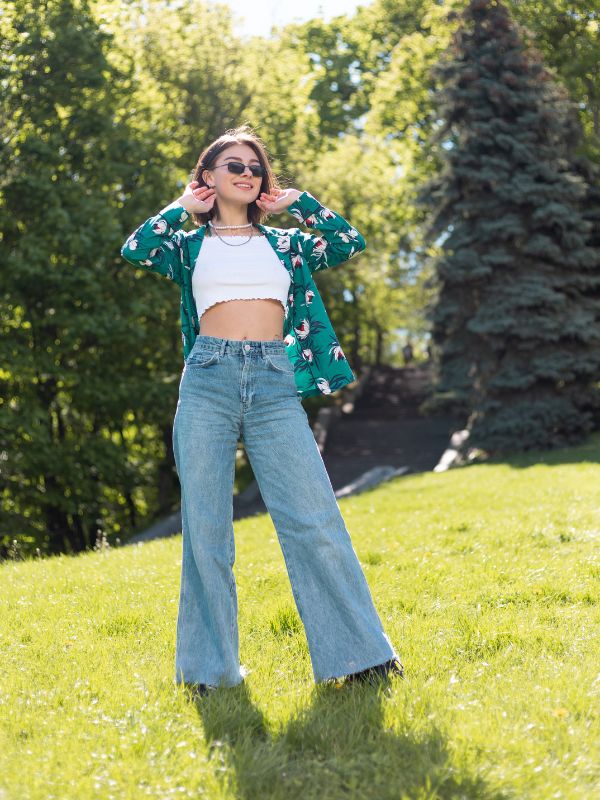 Girl wearing casual outfit with flare jeans, white crop top, and dark green floral shirt standing on grass in sunny day.